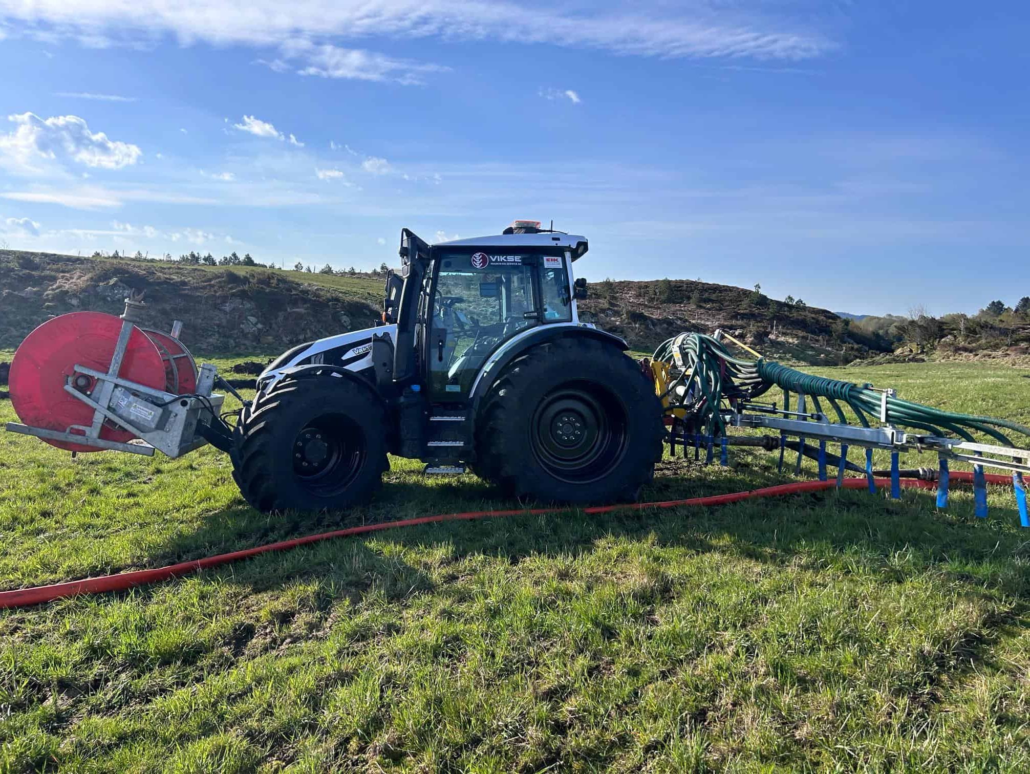 Svart traktor med slangespreder legger ut rød slange på jordet.